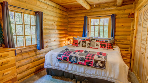 Cozy log cabin bedroom with a bed adorned in a patterned quilt, wooden furniture, and large windows letting in natural light.